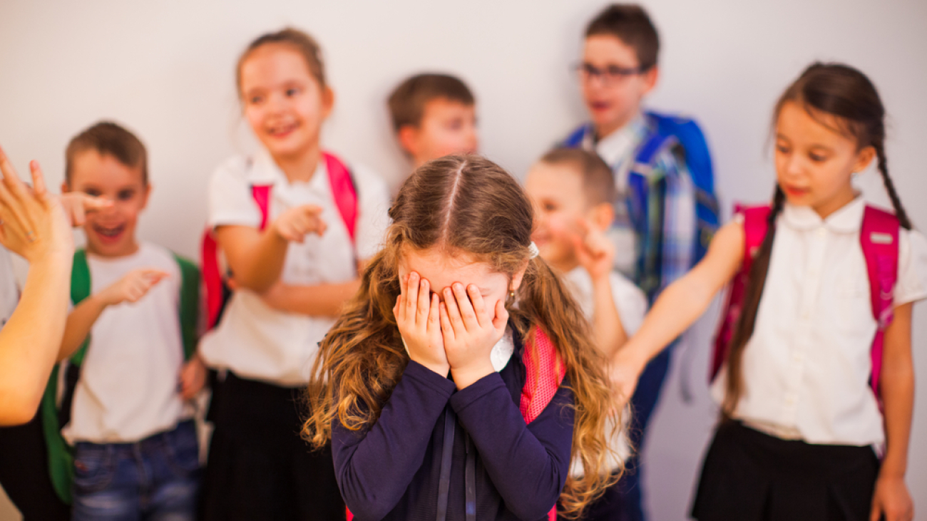 Elementary school girl suffers bullying by classmates. The sad girl covering her face with her hands stands among classmates who point fingers at her