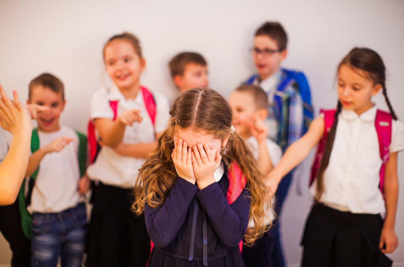 Elementary school girl suffers bullying by classmates. The sad girl covering her face with her hands stands among classmates who point fingers at her