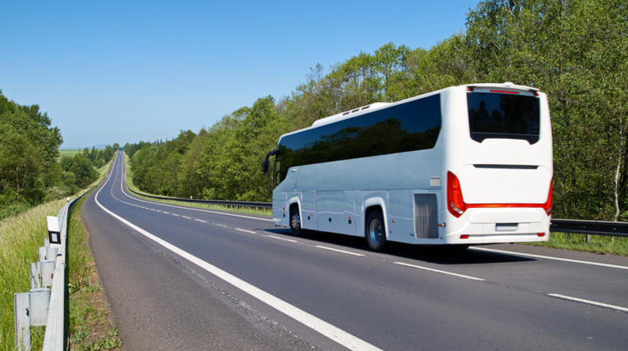 White Bus driving along an empty asphalt road lined with deciduous trees in the countryside